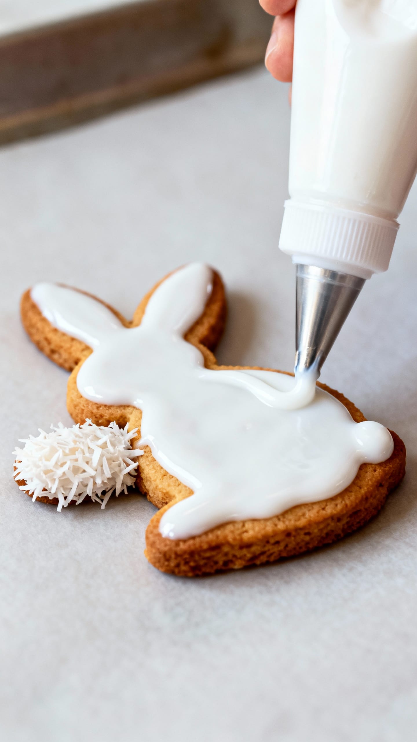 Detail shot: icing bottle piping white glaze, coconut “tail” on bunny cookie